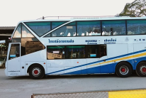 Autocarro branco e azul de dois pisos com letras tailandesas, estacionado num parque pavimentado junto a árvores