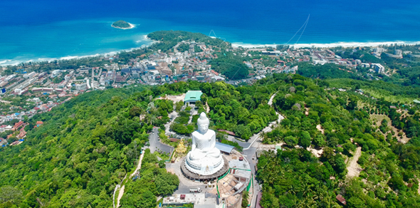 Aerial view of the white Big Buddha statue on a forested hilltop above a coastal town and turquoise sea, Phuket