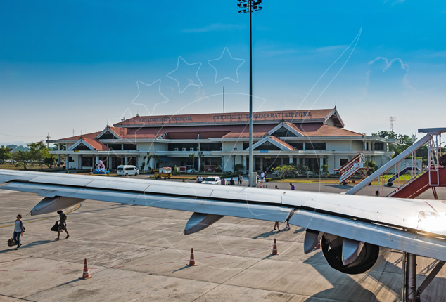 Asa e motor de avião na placa diante de um terminal de aeroporto tropical baixo com telhado vermelho sob céu azul