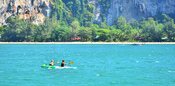 Two people paddle a yellow-green kayak on turquoise sea below limestone cliffs and a tree-lined beach