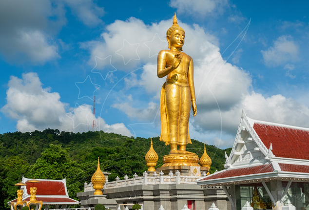 Alta estátua dourada de Buda em pé sobre templo branco de telhado vermelho numa colina arborizada, Hat Yai, Tailândia