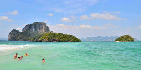 Swimmers in turquoise shallows off a white-sand beach facing limestone karst islands under blue sky, Thailand
