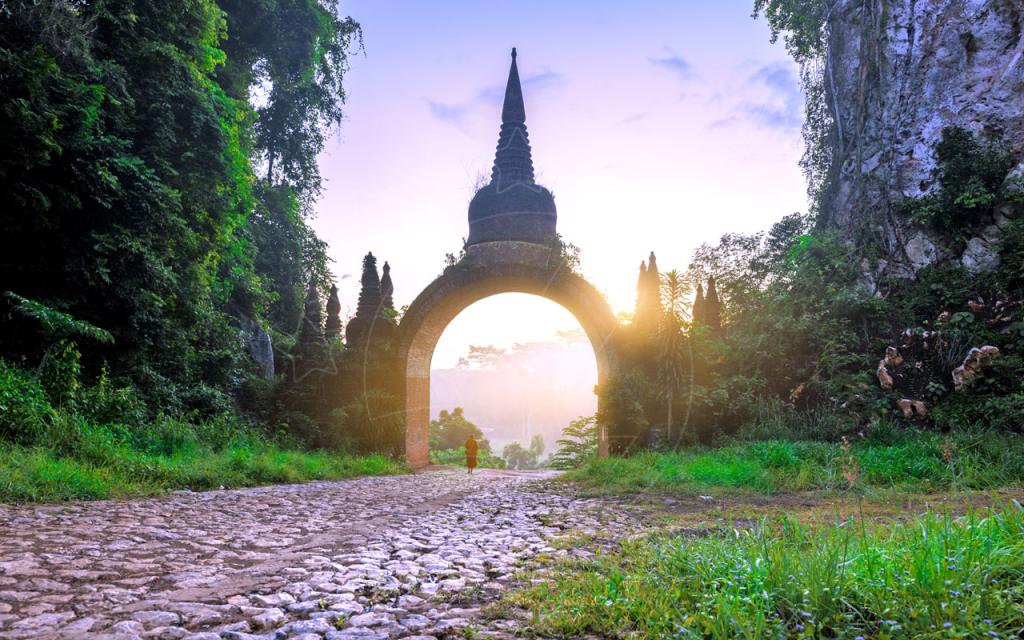 Portal antigo de templo em arco com pináculo em silhueta ao amanhecer, monge a caminhar por trilho de pedra entre falésias verdejantes