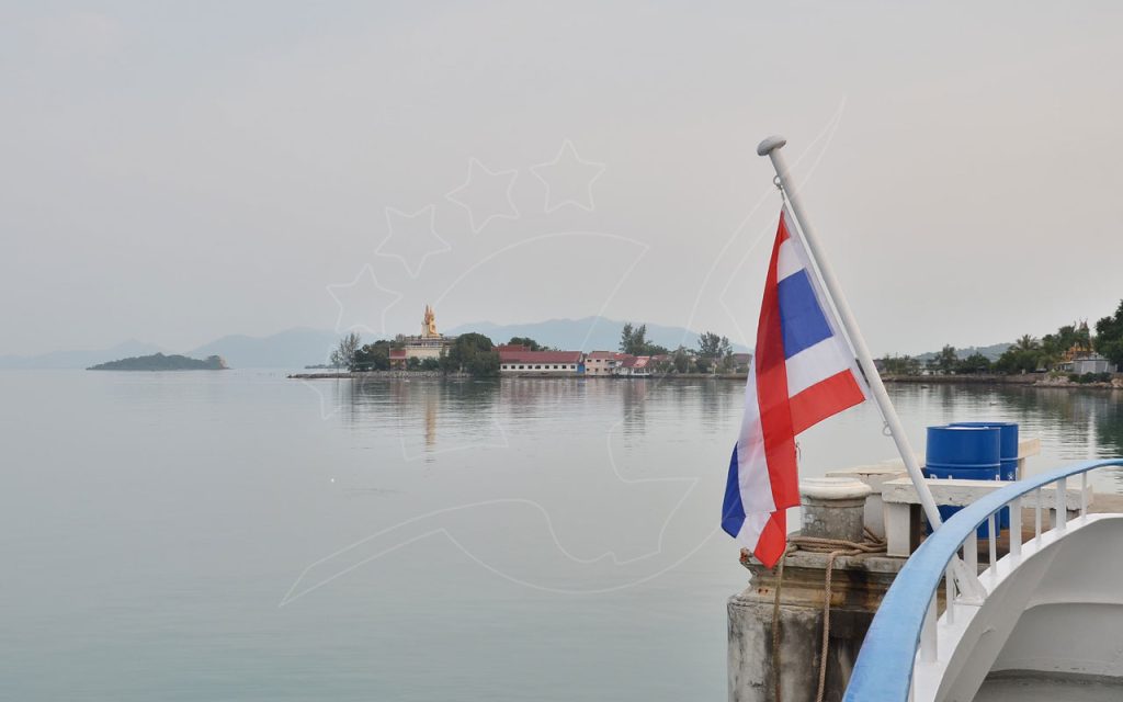 Bandeira da Tailândia na balaustrada de um cais sobre mar calmo e enevoado, com templo e Buda dourado ao longe num promontório
