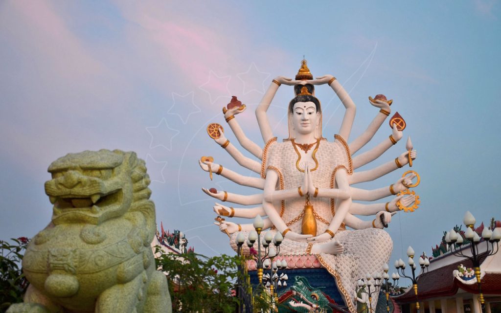 Estátua branca de Guanyin com dezoito braços no templo Wat Plai Laem, Koh Samui, com leão guardião de pedra sob céu pastel