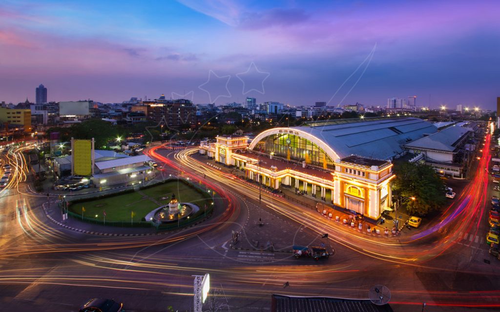 Estação ferroviária de Hua Lamphong iluminada em Banguecoque ao crepúsculo, com rastos de luzes de carros em torno de uma rotunda com fonte