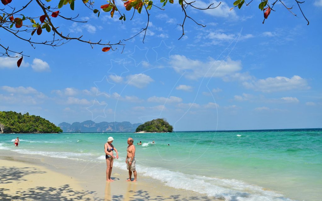 Casal idoso na água turquesa rasa de uma praia de areia branca com ilhas calcárias ao largo, Tailândia