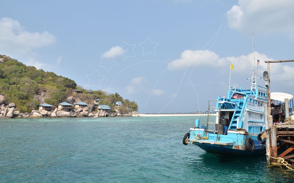 Barco de pesca tailandês de madeira azul atracado num cais gasto junto a baía turquesa com banco de areia e bungalows