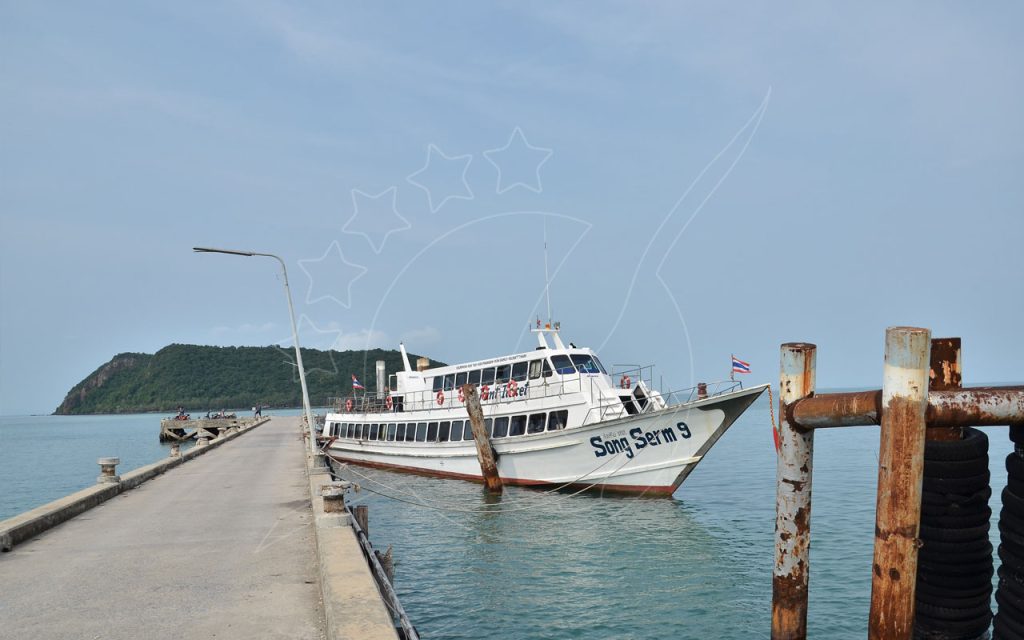 Ferry Songserm atracado num longo cais de betão sob céu azul enevoado, promontório verde de ilha ao fundo