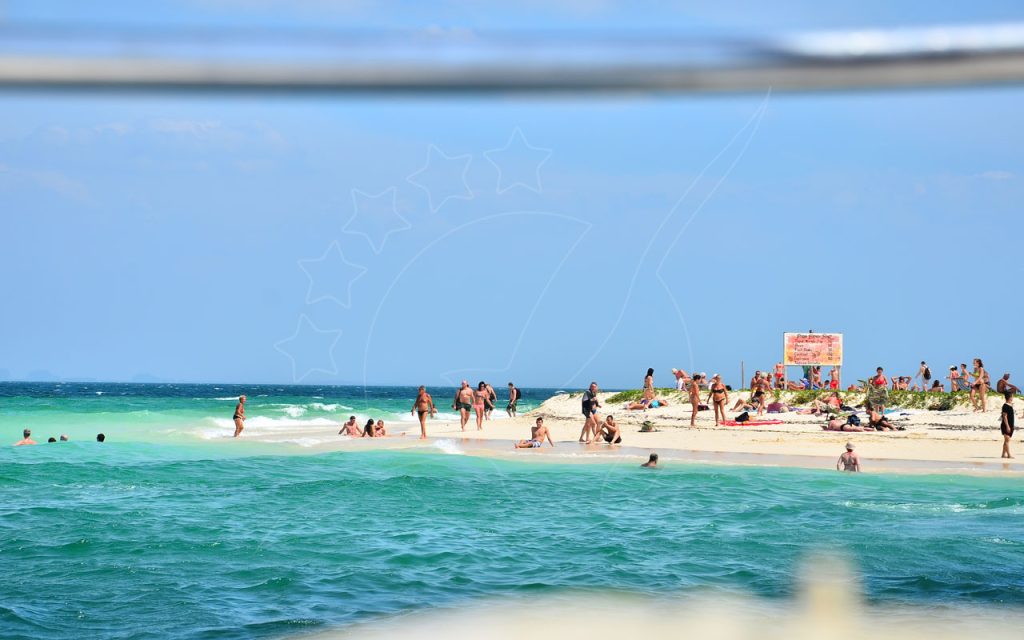 Turistas a apanhar sol e a nadar num banco de areia tropical estreito com mar turquesa e tabela de preços de bebidas