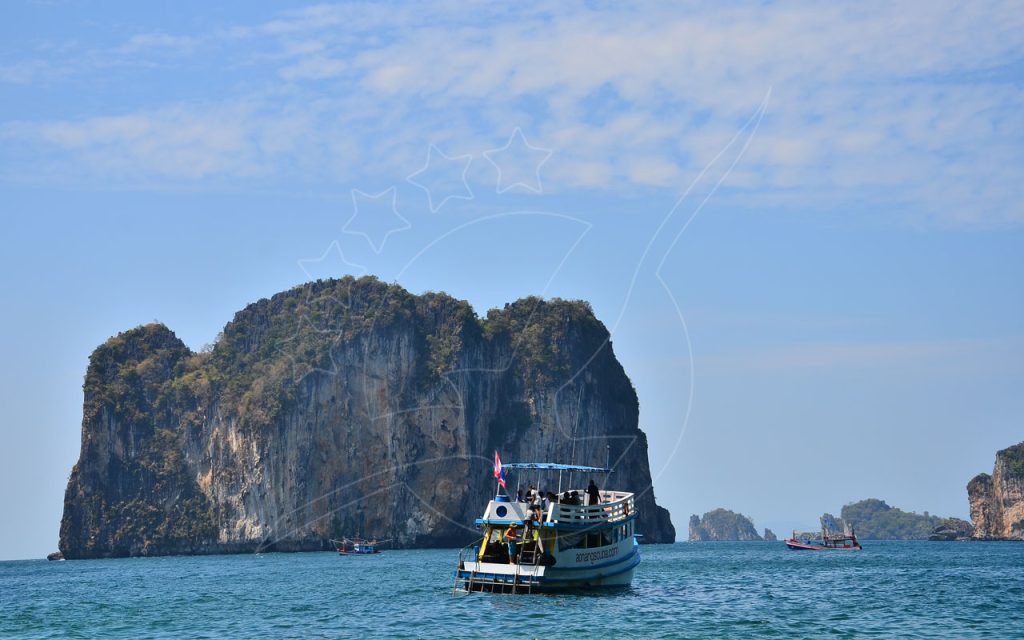 Barco de mergulho de madeira com passageiros ancorado junto a imponente ilha de calcário em mar turquesa calmo