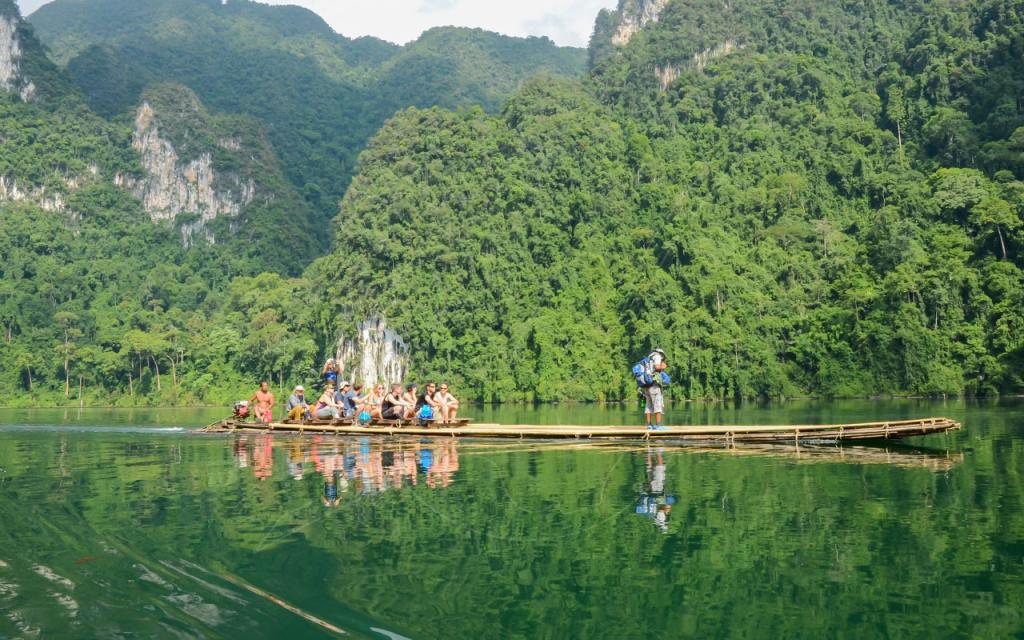Viajantes sentados numa longa jangada de bambu a atravessar um lago verde calmo sob colinas calcárias cobertas de selva