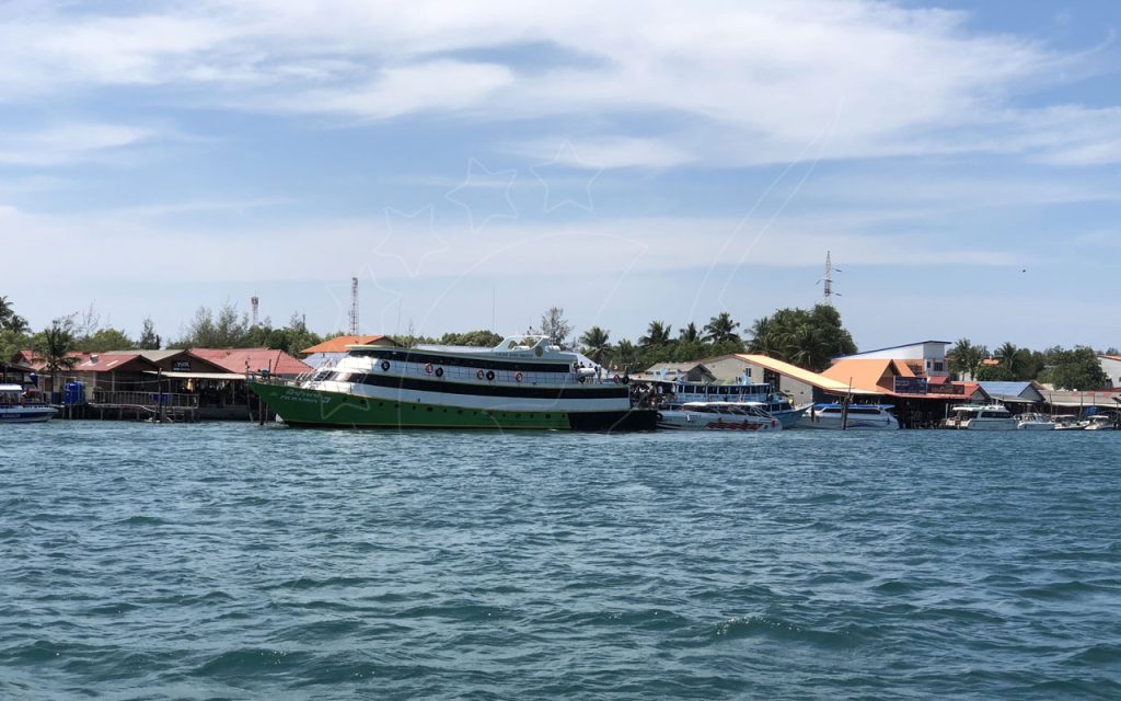 Ferry de passageiros verde e branco atracado num cais com edifícios sobre estacas, visto do mar agitado