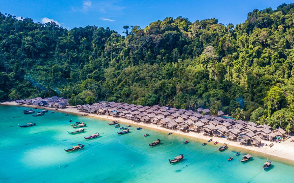 Vista aérea de aldeia palafita com telhados de palha em praia de areia branca e barcos longtail em água turquesa sob colinas de selva