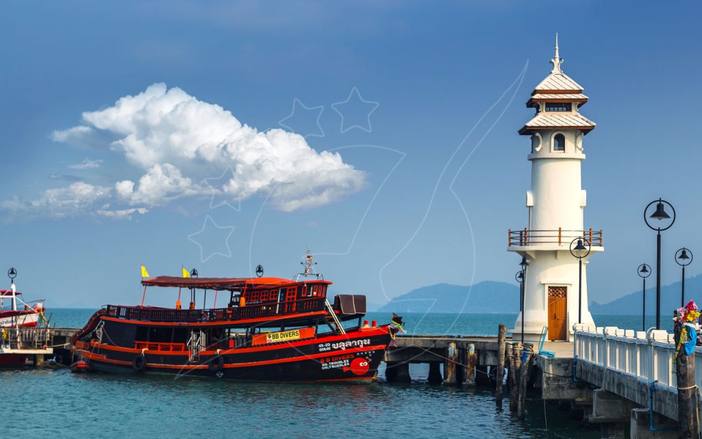 Barco de mergulho de madeira vermelho e preto atracado num cais de betão junto a um farol branco tailandês, céu azul