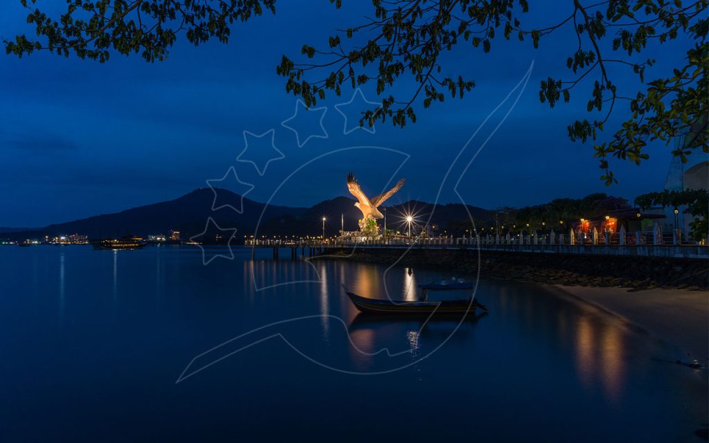 Estátua da águia iluminada na Eagle Square de Langkawi na hora azul, com barco de madeira ancorado em baía calma