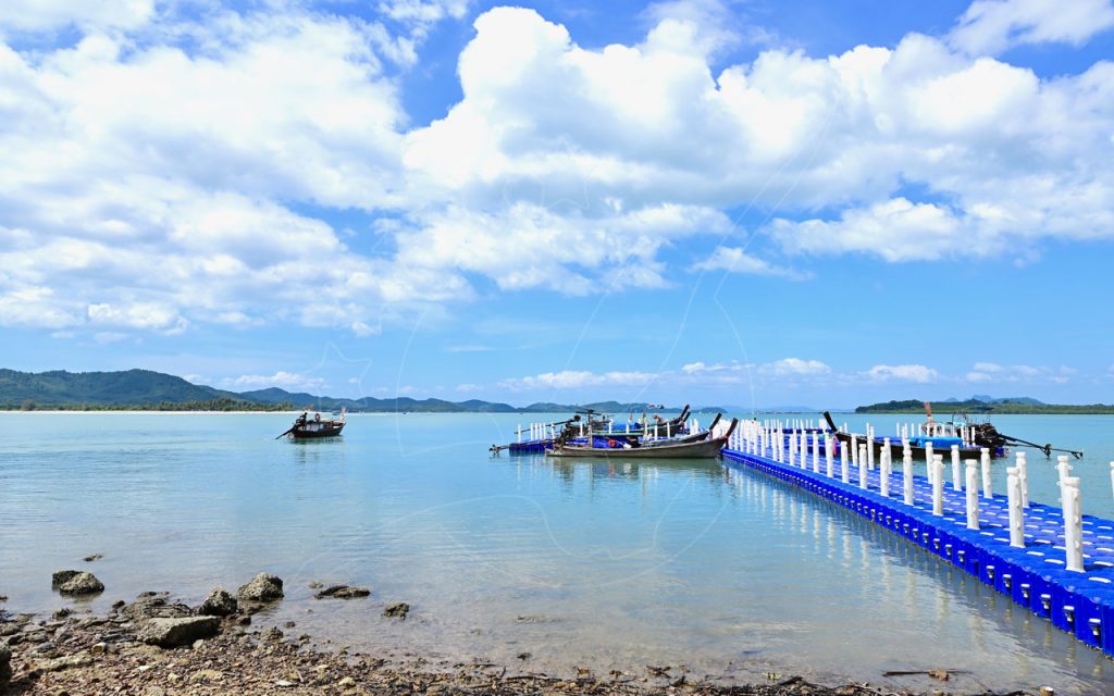 Cais flutuante de pontões de plástico azul com barcos longtail atracados em água turquesa calma sob nuvens cúmulos