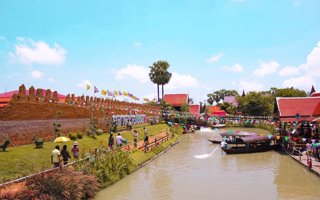 Barcos de cauda longa num canal junto a muro de tijolo e bandeirinhas num mercado flutuante tailandês, com turistas