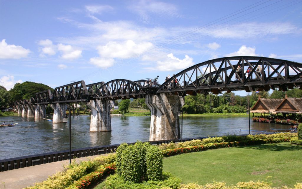 Ponte de treliça de aço negro sobre o rio Kwai, em Kanchanaburi, Tailândia, com turistas a atravessar sob céu azul