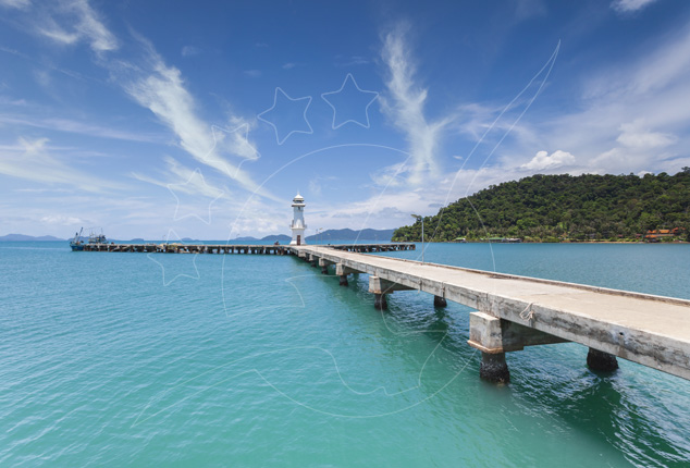 Cais de betão sobre mar turquesa em direção a um pequeno farol branco, com ilha arborizada e céu azul
