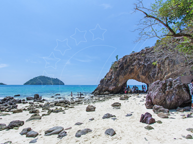 Arco rochoso natural numa praia de areia branca com mar turquesa, rochedos dispersos e ilha verde ao longe sob céu azul