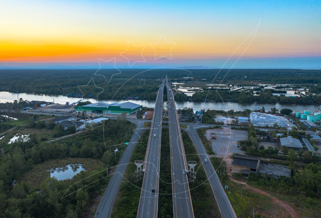 Vista aérea ao anoitecer de uma ponte de autoestrada com separador central sobre um rio largo, junto a armazéns e mangal