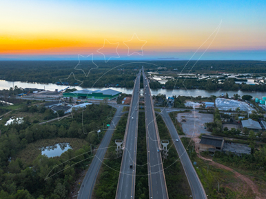 Vista aérea de uma autoestrada com separador central a atravessar um rio em duas pontes ao pôr do sol, com vegetação tropical