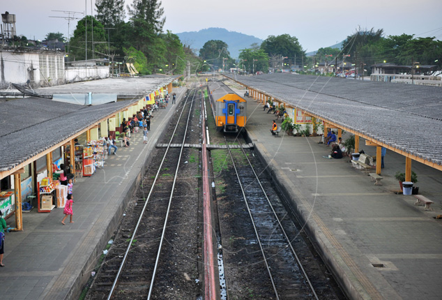 Vista elevada de uma estação ferroviária tailandesa com duas longas plataformas cobertas, carris e uma automotora diesel azul e laranja