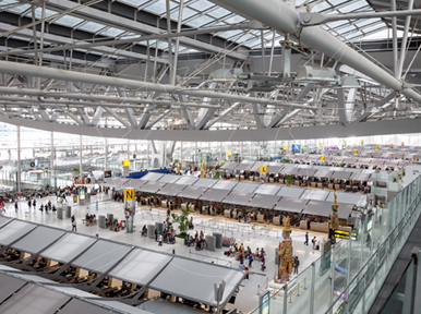 Vista elevada de um terminal aeroportuário moderno com teto de vigas de aço, balcões de check-in e viajantes em baixo