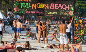 Praia cheia em Koh Phangan com placa pintada à mão "Welcome to Full Moon Beach" e pessoas em fato de banho na areia