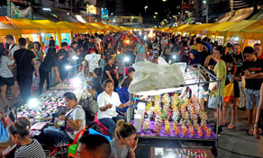 Mercado noturno repleto ao anoitecer com bancas iluminadas, vendedores e clientes a ver lembranças sob toldos