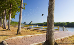 Passeio ribeirinho em tijolo com árvores e guarda branca, rio calmo e colinas calcárias sob céu azul limpo