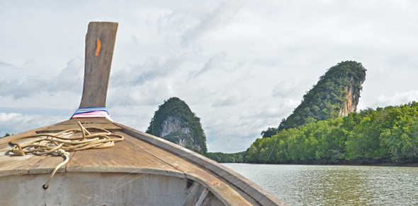 Bow of a wooden longtail boat on a calm river with limestone karst peaks and mangrove forest under cloudy sky