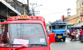 Songthaew vermelho com sinal TAXI no teto estacionado numa rua movimentada tailandesa, songthaew azul e motos atrás
