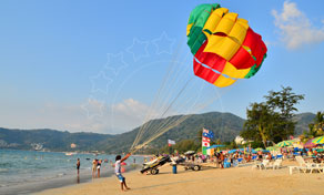 Vela de parapente colorida a encher numa praia tropical movimentada com espreguiçadeiras e promontório arborizado atrás