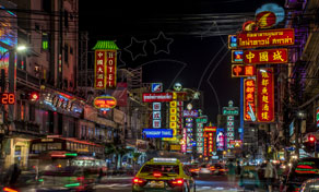 Letreiros de néon em tailandês e chinês iluminam uma rua movimentada da Chinatown de Bangkok à noite, com carros e táxis no trânsito