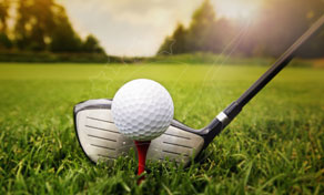 Bola de golfe num tee vermelho junto à cabeça de um driver na relva verde ao sol, com reflexo de lente