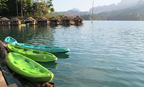 Green kayaks lined along a lake shore with floating bamboo raft houses and limestone hills behind