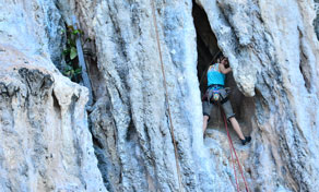 Escalador de camisola de alças azul a subir falésia calcária cinzenta com corda junto a entrada escura de gruta