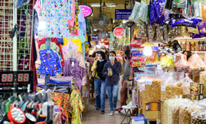 Clientes percorrem um corredor estreito de mercado coberto com bancas de roupa colorida, malas e snacks embalados