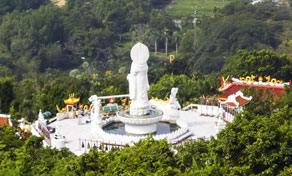 Estátua branca de Guanyin e praça do santuário com pavilhões de telhado vermelho num cume verde arborizado, visto de cima