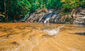 Visitantes a banhar-se numa poça de cascata na selva com água acastanhada e floresta verde densa, Tailândia
