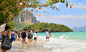 Turistas a caminhar em água turquesa rasa numa praia tropical sob falésias calcárias, entre ramos de árvores