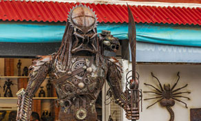 Escultura de sucata em tamanho real estilo Predador com lâmina junto a figura de alienígena numa loja de artesanato
