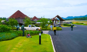 Entrada de resort com pavilhões de telha, jardim relvado, jardineiros a cortar a relva e pickup branca no acesso