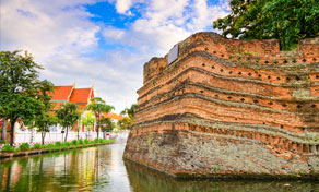 Baluarte de canto em tijolo vermelho envelhecido da antiga muralha de Chiang Mai junto ao fosso, com telhados de templos e palmeiras atrás