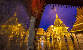 Chedi dourado e pavilhões do templo iluminados ao crepúsculo, refletidos em azulejos molhados, com visitantes desfocados a passar