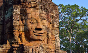 Rosto de pedra esculpido numa torre de arenito do templo Bayon, Angkor, entre árvores verdes e céu azul