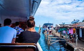 Passageiros sentados no convés de um barco a chegar ao cais de uma aldeia de palafitas com casas coloridas sobre água turquesa