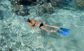 Praticante de snorkeling com barbatanas azuis a flutuar de bruços em água turquesa clara sobre rochas e coral